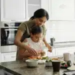 Mother teaching daughter to prepare a healthy meal in a bright, modern kitchen — a heartwarming moment that reflects family traditions, home cooking, and the love behind every recipe.